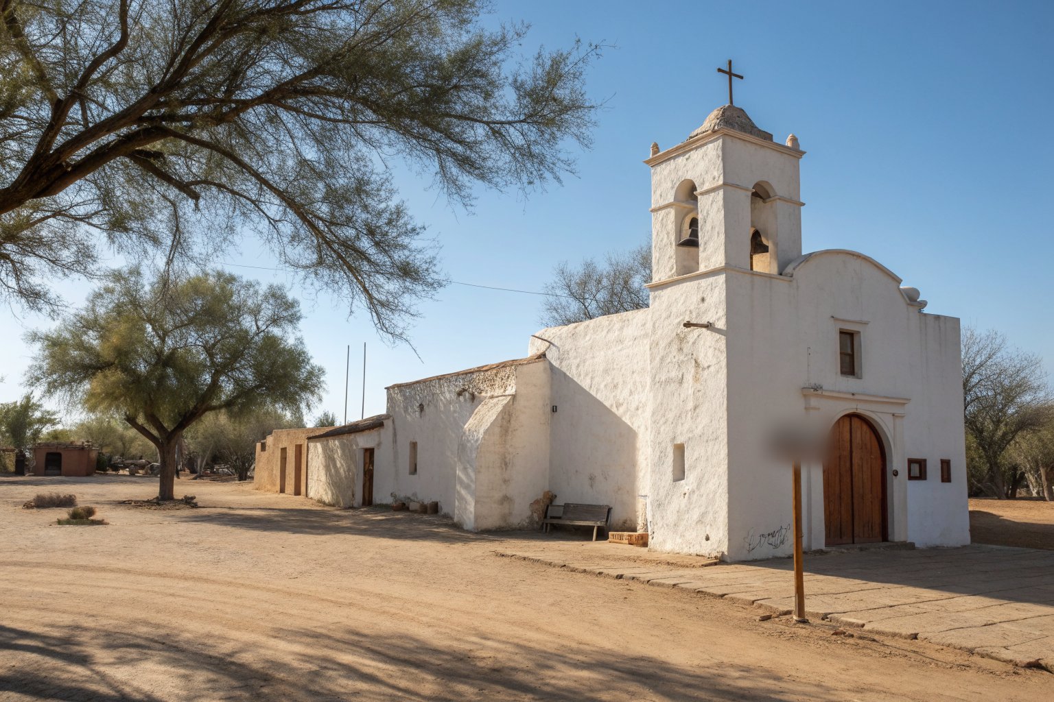 A whitewashed adobe mission church with a bell tower and arched wooden door on a dirt plaza