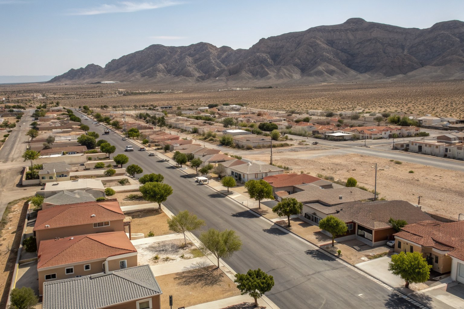 Aerial view of a desert neighborhood of tile- and flat-roofed homes along a wide street, with bare mountains beyond